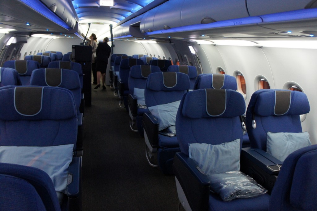 The image shows the interior of an airplane cabin, specifically the business class section. The seats are arranged in a 2-2 configuration, with wide, blue cushioned seats that have headrests and armrests. Each seat has a pillow and a blanket placed on it. The cabin is well-lit with blue ambient lighting on the ceiling. There are a few passengers and flight attendants visible in the background, near the front of the cabin. The windows along the side of the plane are visible, and the overall atmosphere appears clean and organized.