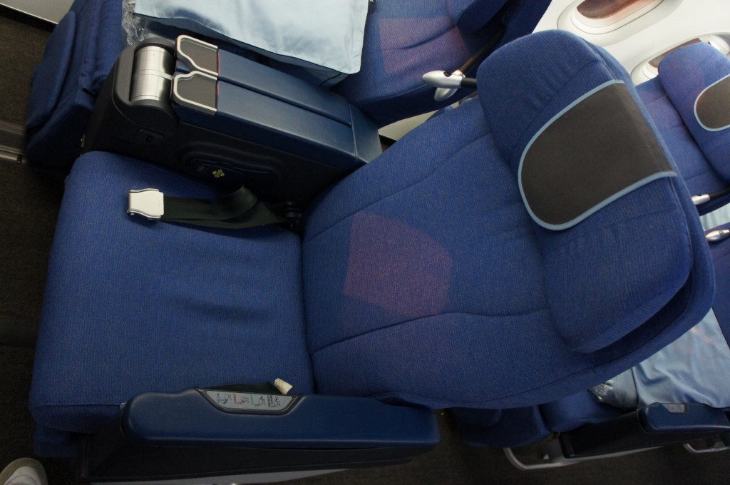 The image shows a blue airplane seat in a reclined position. The seat has a headrest with a dark-colored cushion, an armrest with a control panel, and a seatbelt. The seat is part of a row of similar seats, and there is a window visible in the background. The overall setting appears to be the interior of an airplane.