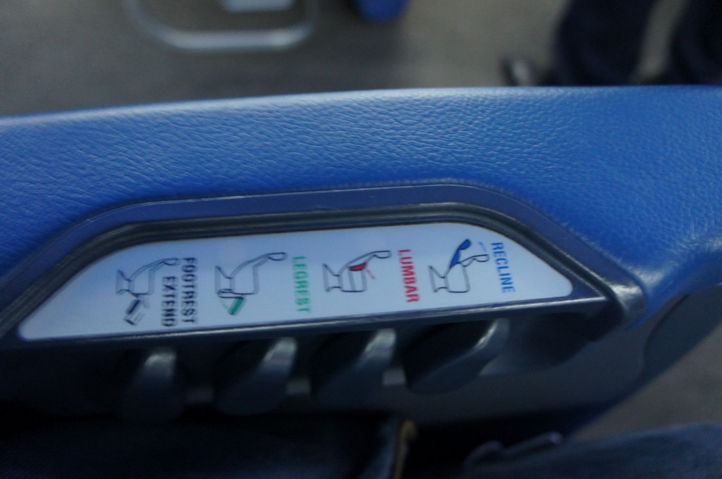 The image shows a close-up of a control panel on the armrest of a seat, likely in an airplane or a bus. The panel has four labeled buttons with corresponding icons indicating their functions: "Footrest Extend," "Legrest," "Lumbar," and "Recline." The armrest is covered in blue material.