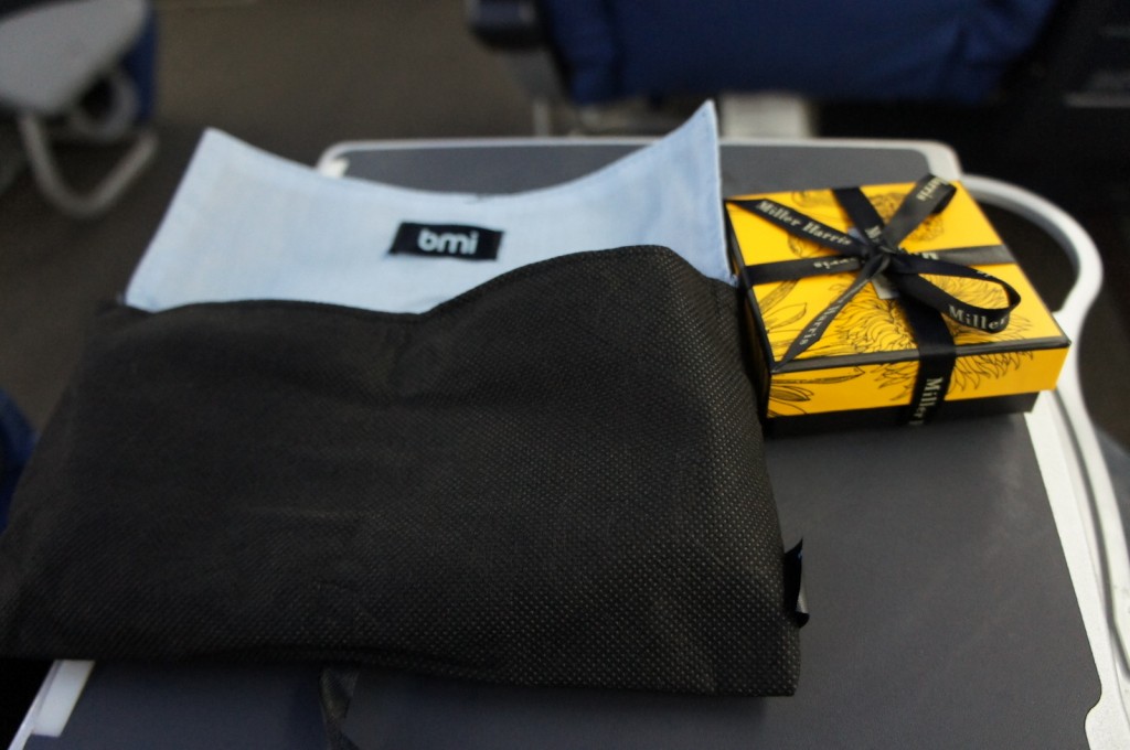 The image shows a tray table on an airplane. On the tray table, there is a black fabric pouch with a light blue interior and a label that reads "bmi." Next to the pouch, there is a yellow box with black ribbon tied around it, featuring the text "Miller Harris" on the ribbon. The background includes parts of airplane seats and other tray tables.