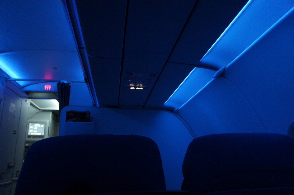 Alt text: The interior of an airplane cabin is dimly lit with blue lighting. The image shows the ceiling, overhead compartments, and part of the seats. The entrance to the galley area is visible in the background, with a faintly illuminated exit sign above it.