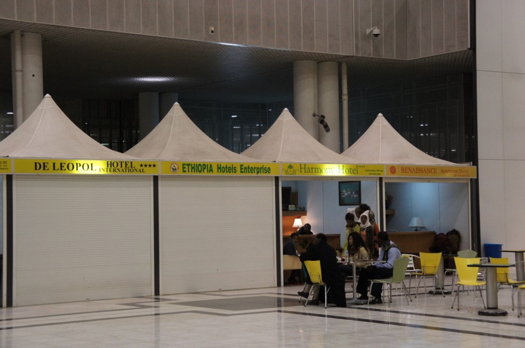 The image shows a row of hotel service booths in an indoor setting, likely an airport or a convention center. The booths have white tent-like structures with yellow signage displaying the names of different hotels: "DE LEOPOL HOTEL INTERNATIONAL," "ETHIOPIA Hotels Enterprise," "Harmony Hotel," and "RENAISSANCE Apartment Hotel." The first and last booths are closed with shutters down, while the middle two booths are open and staffed. In front of the booths, there are several people sitting at tables with yellow chairs, engaged in conversation or working on laptops. The area is well-lit and has a modern design with a tiled floor.