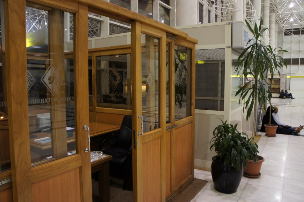 The image shows a small wooden office booth with glass windows and doors, located inside a large, open indoor space. The booth has a sign on the glass that reads "Sheraton Addis." Inside the booth, there is a black office chair and a desk with some items on it. To the right of the booth, there are two potted plants. In the background, there are people sitting on the floor against the wall, and the area appears to be well-lit with high ceilings and large windows.
