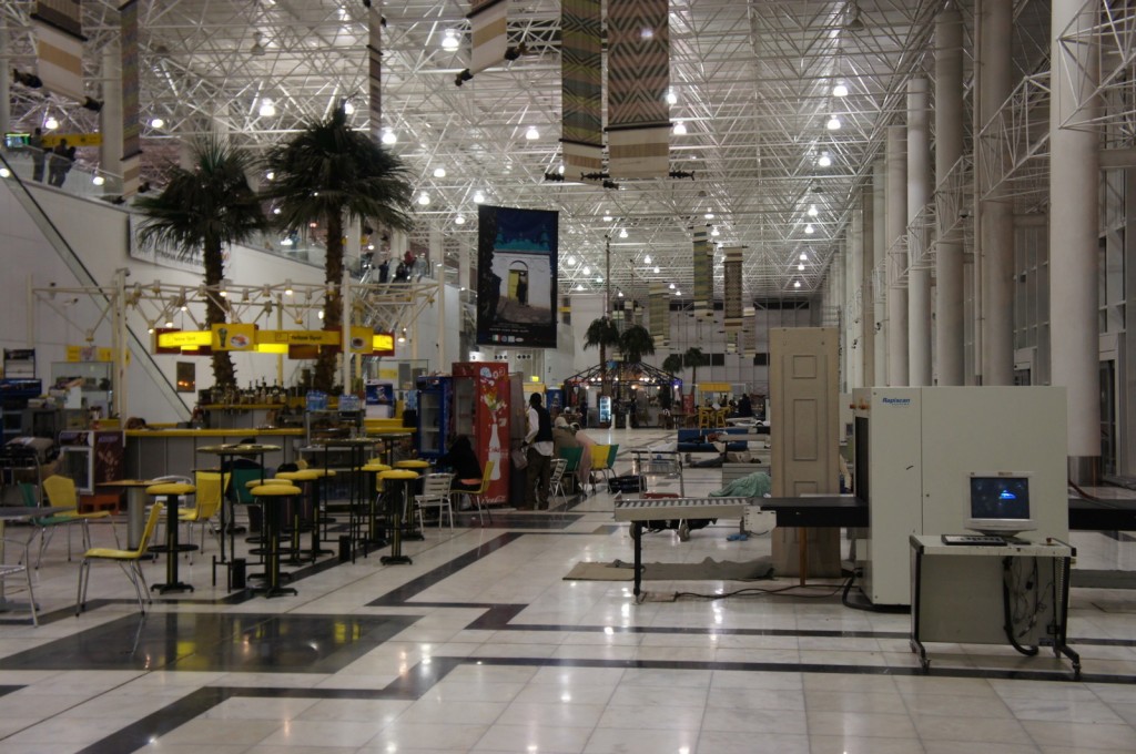The image shows the interior of an airport terminal. The terminal is spacious with high ceilings and a modern design featuring a lot of white and metallic elements. There are several palm trees placed throughout the area, adding a touch of greenery. In the foreground, there are tables and chairs, some of which are yellow, arranged in a seating area. To the right, there is a security scanning machine with a conveyor belt. In the background, there are various kiosks and shops, including a yellow sign for a food or beverage stand. The terminal is well-lit with numerous lights hanging from the ceiling, and there are a few people visible in the distance.