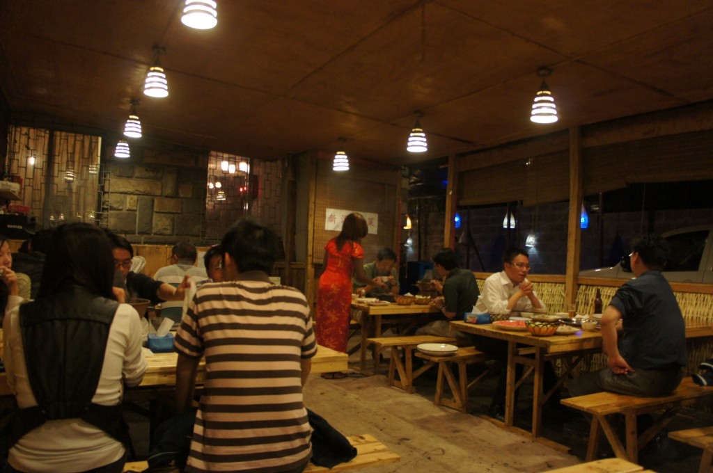 The image shows the interior of a cozy restaurant with wooden furniture and warm lighting. Several groups of people are seated at wooden tables, engaged in conversation and enjoying their meals. The restaurant has a rustic ambiance with hanging pendant lights and a wooden ceiling. A person in a red dress is standing and appears to be serving or interacting with the diners. The overall atmosphere is lively and inviting.