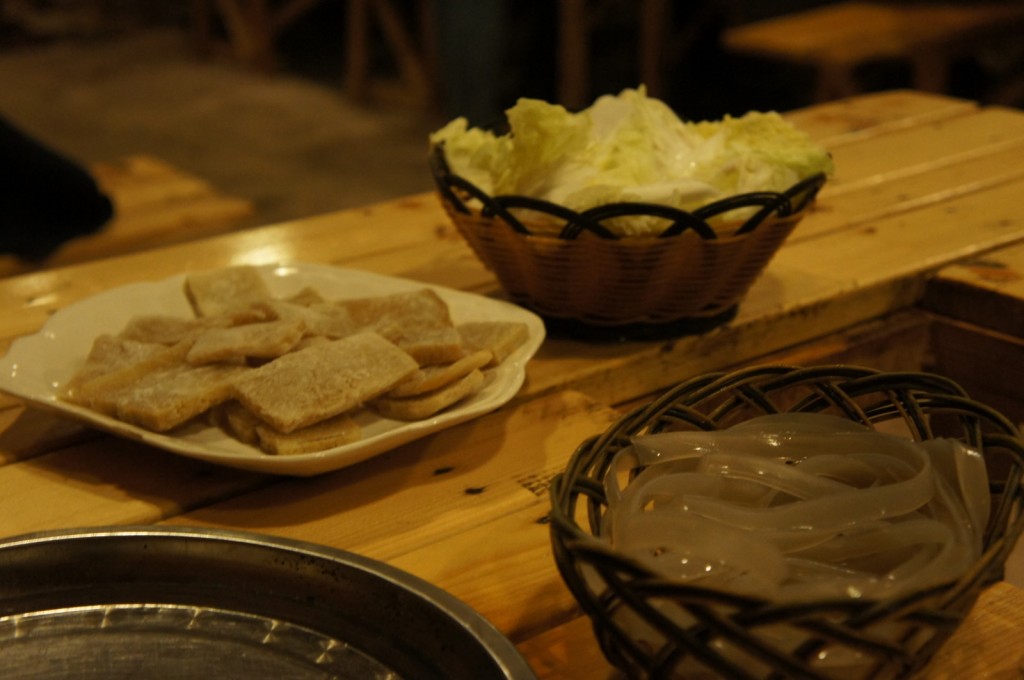 A wooden table with three different food items. On the left, there is a white plate with rectangular pieces of food, possibly tofu or a similar ingredient. In the middle, there is a basket containing leafy greens, likely cabbage. On the right, there is another basket with translucent, noodle-like strips. The setting appears to be a rustic or casual dining environment.