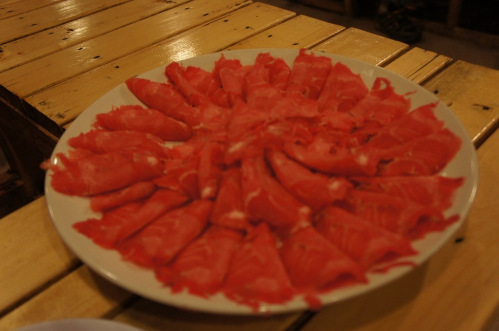 A large white plate filled with thinly sliced raw meat, arranged in a circular pattern. The plate is placed on a wooden table.