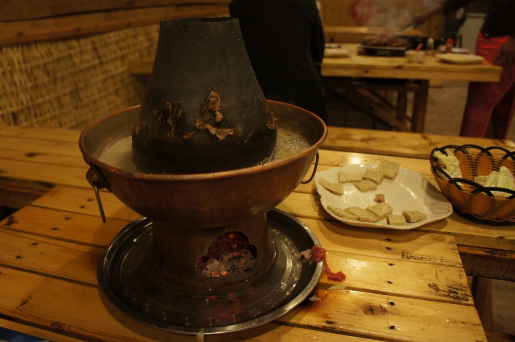 The image shows a traditional Chinese hot pot setup on a wooden table. The hot pot has a central chimney and is filled with boiling broth. There are plates around the hot pot, one with pieces of tofu and another with vegetables. The table is set in a rustic environment, possibly a restaurant, with wooden benches and a woven wall in the background. There are people in the background, but they are not the main focus of the image.