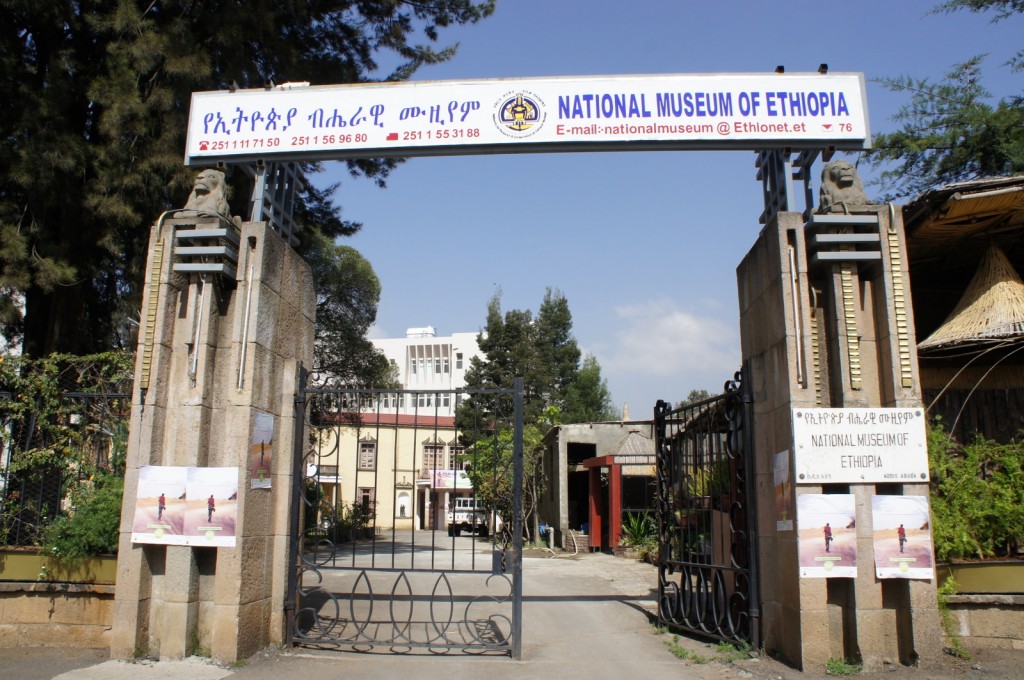 The image shows the entrance gate of the National Museum of Ethiopia. The gate is flanked by two stone pillars, each topped with a lion statue. Above the gate is a large sign with text in both Amharic and English, reading "National Museum of Ethiopia" along with contact information. The entrance is open, revealing a pathway leading to the museum building in the background, surrounded by trees and greenery. There are also posters displayed on the stone pillars near the entrance.
