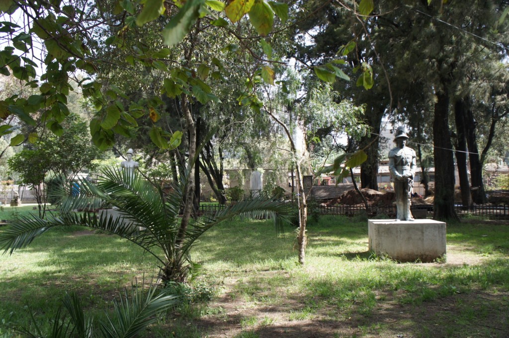 The image shows a serene park with lush green grass and various trees providing shade. In the foreground, there is a small palm tree. Towards the right side of the image, there is a statue of a person standing on a concrete pedestal. The statue appears to be of a soldier or a person in uniform. The background includes more trees and some structures that are partially visible through the foliage. The overall atmosphere is peaceful and green.