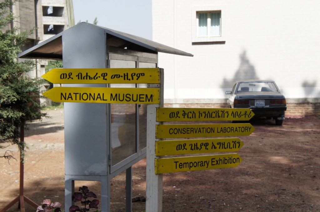 The image shows a signpost with multiple yellow directional signs in both Amharic and English. The signs point to different locations within a museum complex. The directions include "National Museum," "Conservation Laboratory," and "Temporary Exhibition." The signpost is situated outdoors, with a building and a parked car visible in the background. There are also some plants at the base of the signpost.