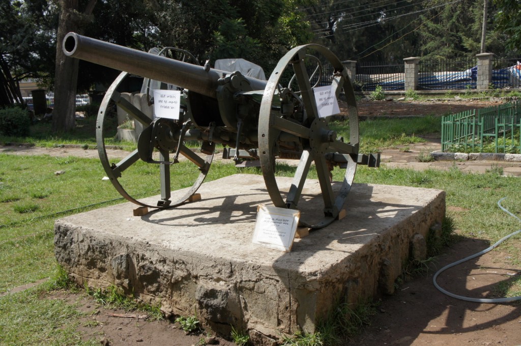 The image shows an old artillery cannon displayed outdoors on a concrete pedestal. The cannon has large spoked wheels and is situated in a grassy area with trees and a fence in the background. There are informational signs attached to the cannon and placed on the pedestal, providing details about the artifact. The setting appears to be a park or a museum garden.