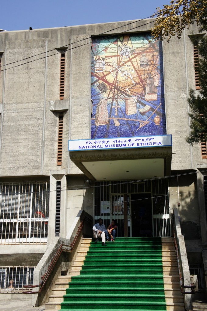 The image shows the entrance to the National Museum of Ethiopia. The building is made of concrete and has a large, colorful mosaic above the entrance depicting various historical and cultural elements. There is a sign in both Amharic and English that reads "National Museum of Ethiopia" above the entrance. A set of green-carpeted stairs leads up to the entrance, and two people are sitting on the steps. The building has barred windows on either side of the entrance. A tree is visible on the right side of the image.
