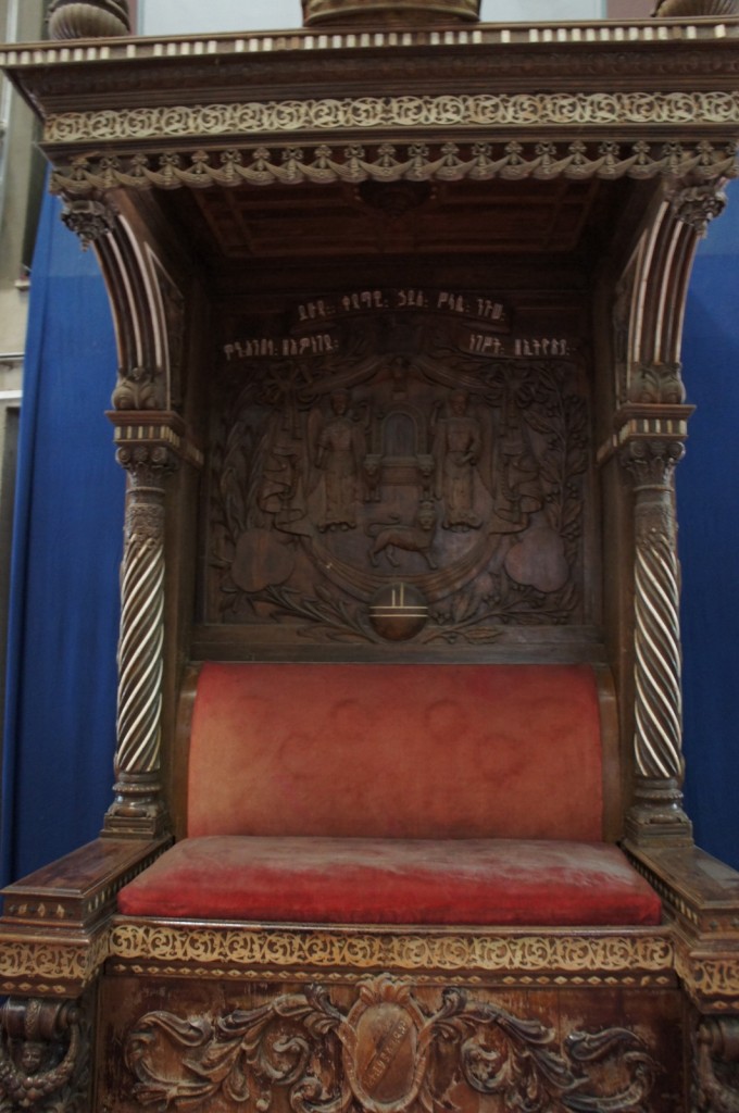 The image shows an ornate wooden throne with intricate carvings and a red cushioned seat and backrest. The throne features detailed woodwork, including twisted columns on either side and elaborate patterns along the top and bottom edges. The backrest has a carved scene with figures and text in a script that appears to be ancient or decorative. The throne is set against a blue background.