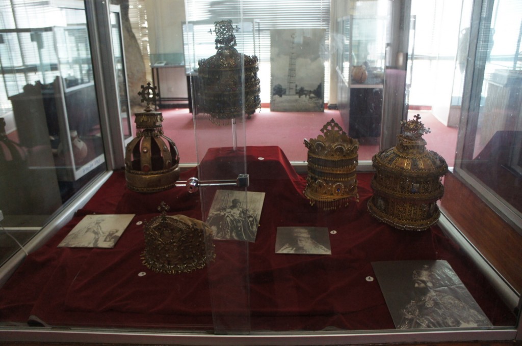 The image shows a display case in a museum containing several ornate crowns and headpieces. The crowns are elaborately decorated with intricate designs and embellishments. The display also includes a few black and white photographs of individuals wearing similar crowns, placed on a red velvet cloth. The background of the image shows other museum exhibits and artifacts.
