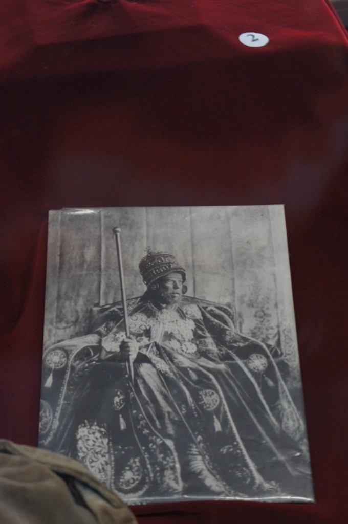 A black and white photograph of a person dressed in elaborate, traditional attire, including a richly decorated robe and a crown. The person is seated on a throne and holding a long staff. The photograph is displayed on a red cloth with a small white label marked with the number "2" in the upper right corner.