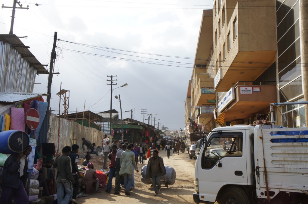 A busy street scene in an urban area. On the left side, there are makeshift stalls and vendors selling various goods, with people walking and shopping. On the right side, there are multi-story buildings with businesses and signs. A white truck is parked on the right side of the street. The street is bustling with pedestrians and activity, and power lines are visible overhead. The sky is cloudy.