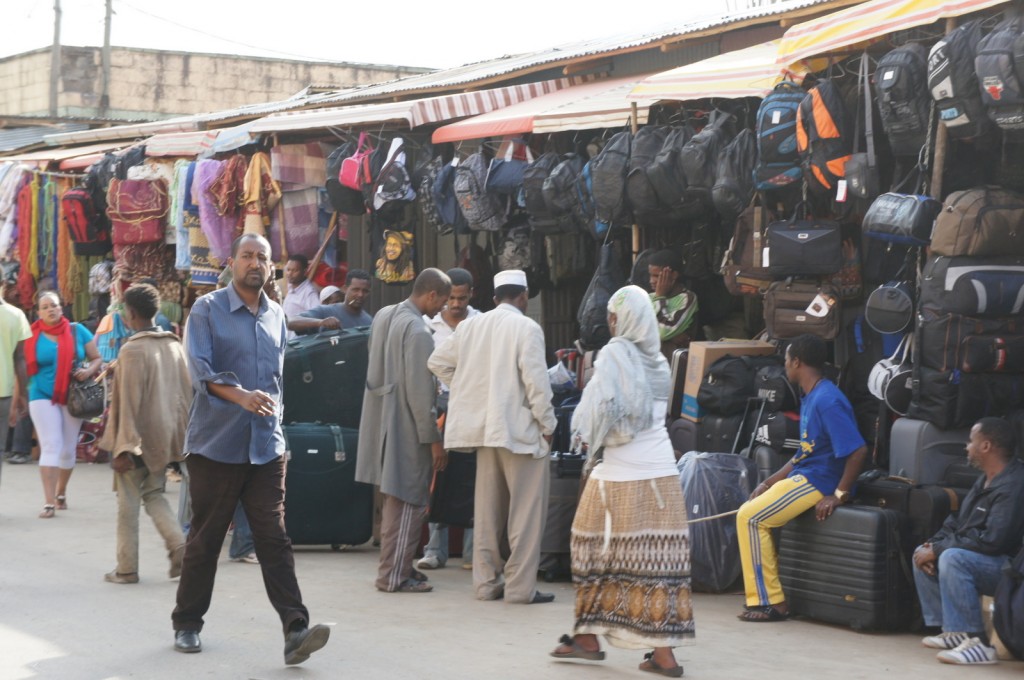 A bustling outdoor market scene with several people walking and interacting. The market stalls are filled with various goods, including bags, suitcases, and colorful textiles. Some people are browsing the items, while others are engaged in conversation. The atmosphere appears lively and busy.