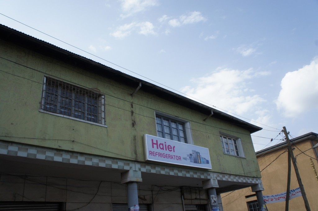 The image shows the exterior of a two-story building with a greenish facade. The building has barred windows on the upper floor and a sign that reads "Haier REFRIGERATOR" with images of refrigerators on it. The sky is partly cloudy, and there are power lines and a utility pole visible on the right side of the image.