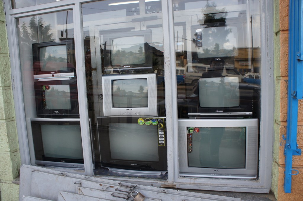The image shows a storefront window displaying a variety of old CRT (cathode-ray tube) televisions. The TVs are stacked and arranged in rows, with different brands and sizes visible. The reflection of the street and surrounding area can be seen in the glass of the window. The window frame is painted gray, and part of the building's exterior, which is painted in light green and orange, is also visible.