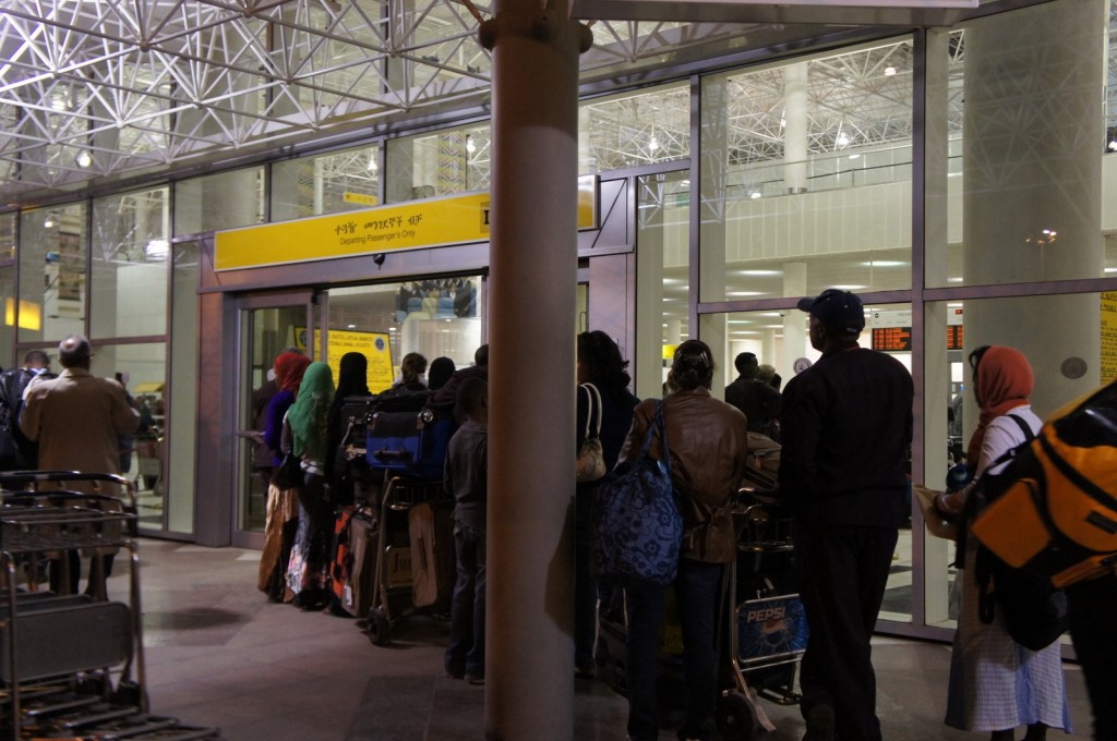 The image shows a group of people standing in line at an airport entrance marked "Departing Passengers Only." The entrance has a yellow sign with text in multiple languages. The people in line are carrying luggage and some are using luggage carts. The interior of the airport is visible through the glass windows, showing a modern design with high ceilings and bright lighting.