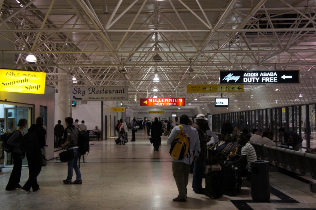 The image shows the interior of an airport terminal with several people walking and sitting. There are various signs indicating different facilities, including a "Souvenir Shop" with a yellow sign on the left, a "Sky Restaurant" sign in the middle, and a "Millennium Duty Free" sign in red further down the corridor. On the right side, there is a sign for "Addis Ababa Duty Free." The terminal has a high ceiling with a lattice of white beams and is well-lit. People are carrying luggage and some are seated, waiting.