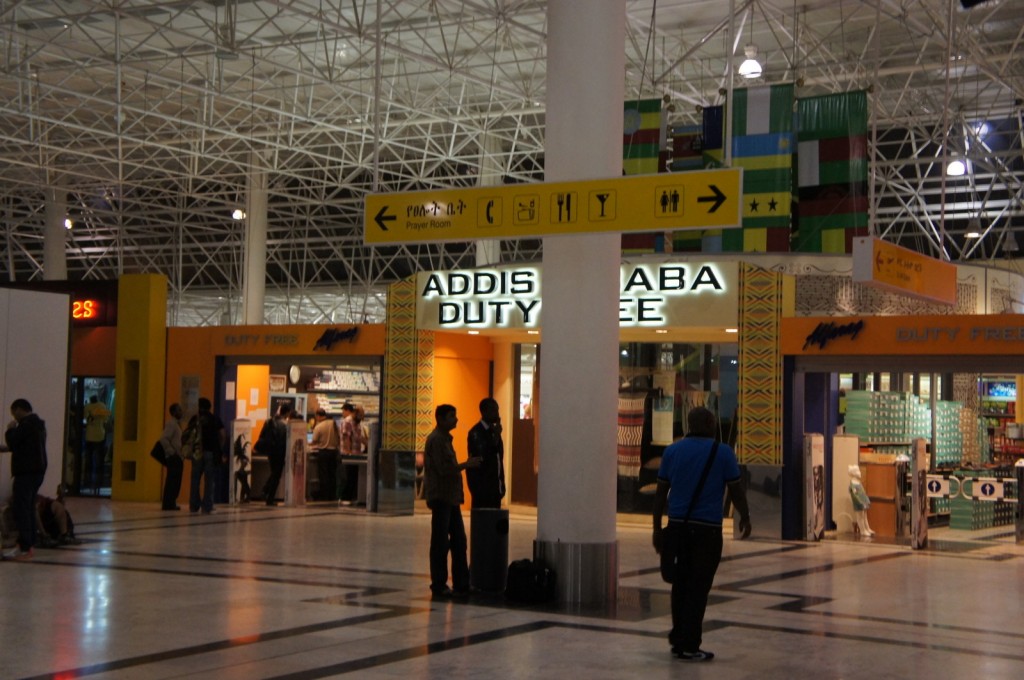 The image shows the interior of an airport terminal with a duty-free shop named "Addis Ababa Duty Free." Several people are seen walking or standing around the area, some with luggage. The ceiling has a complex metal framework, and there are various signs indicating directions to different facilities such as a prayer room, restrooms, and dining areas. The duty-free shop has a bright yellow and orange color scheme, and there are flags hanging from the ceiling in the background.