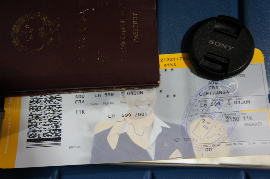 The image shows a close-up of a traveler's items, including a passport, a Lufthansa boarding pass, and a Sony camera lens cap. The boarding pass indicates a flight from Addis Ababa (ADD) to Frankfurt (FRA) on June 4th, with the seat number 11K. The passport is partially covering the boarding pass, and the lens cap is placed on the right side of the boarding pass.
