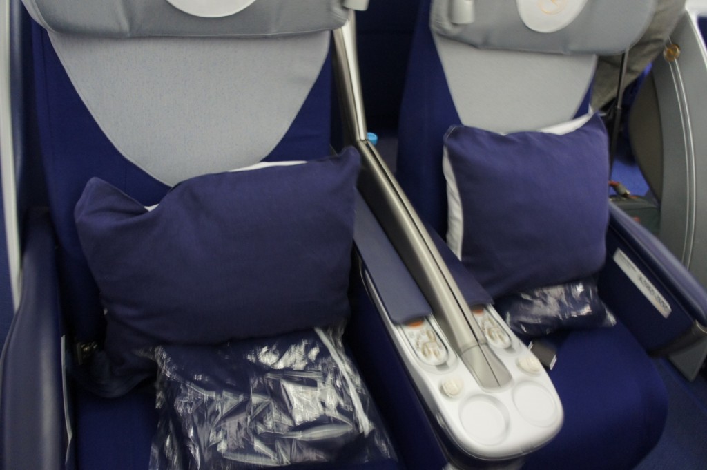 The image shows two adjacent airplane seats in a business or first-class cabin. The seats are upholstered in blue fabric and each has a large, dark blue pillow with a white stripe on the side. There are also packaged blankets or pillows placed on the seats. The seats have a shared armrest with built-in controls and cup holders. The headrests have a light gray cover with a logo on them.