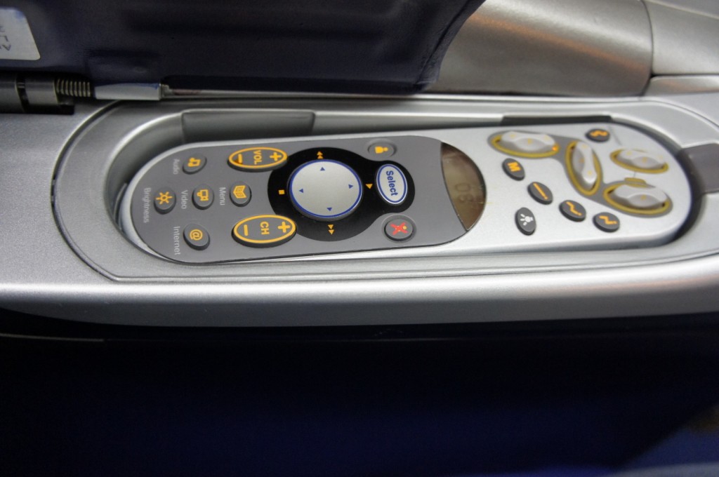 The image shows a close-up of an in-flight entertainment system remote control embedded in the armrest of an airplane seat. The remote has various buttons, including volume control, channel selection, and navigation buttons, as well as buttons for audio, video, brightness, and internet. The remote is housed in a recessed compartment with a cover that can be closed.