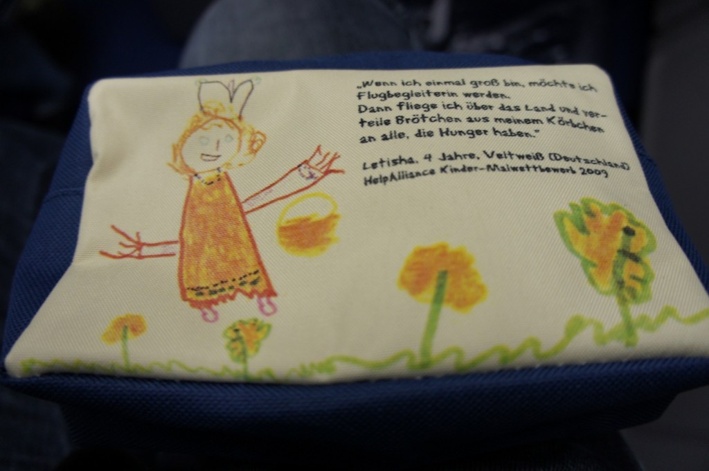 Alt text: A fabric item, possibly a pouch or bag, features a child's drawing and handwritten text. The drawing depicts a person with a smiling face, wearing a dress, and holding a yellow object. There are also flowers and grass drawn at the bottom. The handwritten text is in German and includes the name "Letisha" and mentions "4 Jahre" (4 years old) and "Deutschland" (Germany). The text also references "HelpAlliance Kinder-Malwettbewerb 2009" (HelpAlliance Children's Drawing Competition 2009).