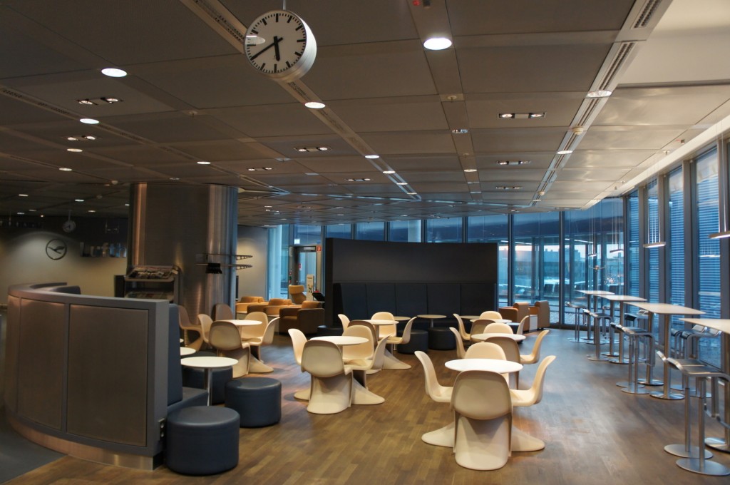 The image shows a modern, spacious lounge area with a variety of seating options. There are several round tables with white chairs, some high tables with bar stools, and a few cushioned seating areas. The floor is wooden, and the ceiling has a grid-like pattern with recessed lighting. Large windows on one side of the room allow natural light to enter, and there are clocks on the walls and ceiling. The overall design is sleek and contemporary, suggesting a comfortable and functional space, possibly in an airport or office building.
