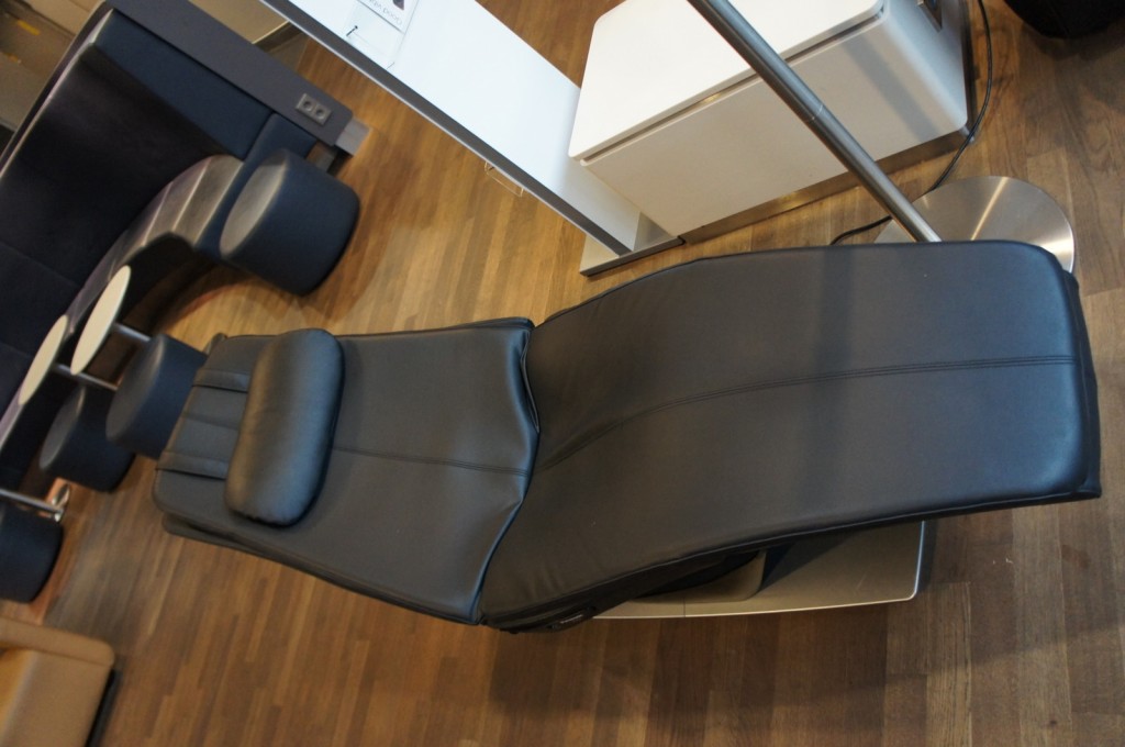 The image shows a modern, black leather reclining chair with a headrest, situated in a room with wooden flooring. In the background, there are other pieces of furniture, including a curved bench with small round tables and stools. There is also a white structure, possibly a partition or a piece of furniture, and a metallic floor lamp. The setting appears to be a lounge or waiting area.