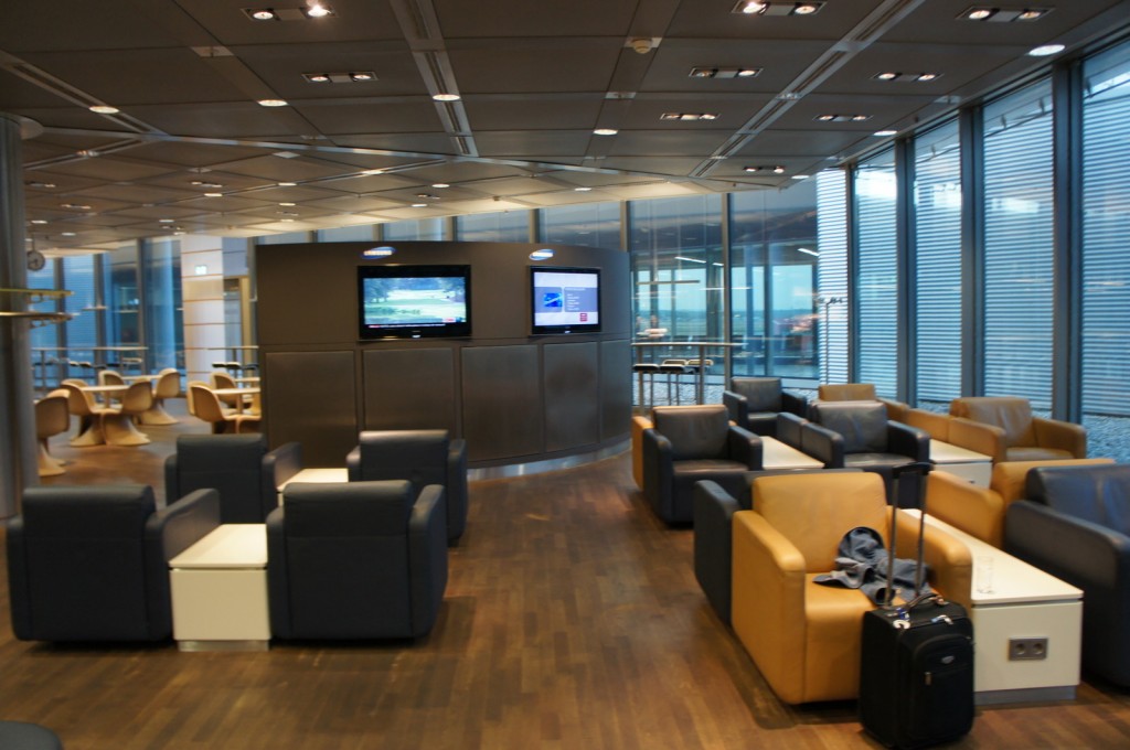 The image shows a modern airport lounge with comfortable seating arrangements. There are several armchairs in shades of blue and beige, each paired with a small table. In the center of the lounge, there are two wall-mounted televisions displaying different channels. The lounge has large windows on one side, allowing natural light to fill the space. In the background, there are more seating areas with round tables and chairs. A piece of luggage and a jacket are placed on one of the armchairs in the foreground. The overall atmosphere is clean and well-lit, providing a comfortable waiting area for travelers.