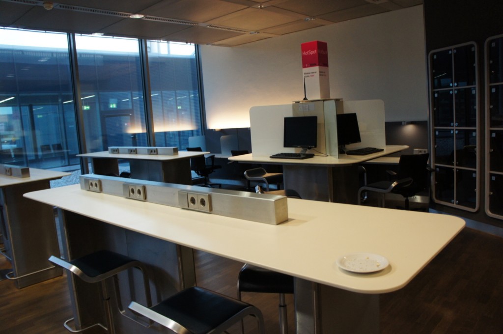 The image shows a modern, well-lit workspace with several long tables and high stools. Each table has multiple power outlets installed on top. In the background, there are computer workstations with chairs, and a sign labeled "HotSpot" is visible above one of the workstations. The room has large windows allowing natural light to enter, and there are lockers on the right side of the image. The floor appears to be made of wood, and the overall design is sleek and functional.