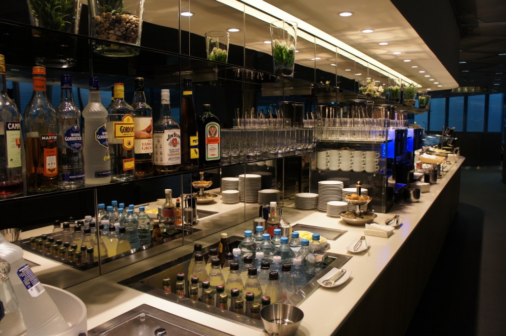 The image shows a well-stocked bar area with a variety of liquor bottles, including brands like Campari, Martini, and Jim Beam, displayed on shelves. Below the shelves, there are numerous glasses neatly arranged. The counter features an assortment of small bottled beverages, water bottles, and various condiments. There are also stacks of plates, cups, and saucers, along with a coffee machine and some snacks. The bar area is well-lit with overhead lighting, and there are decorative plants placed on the shelves.