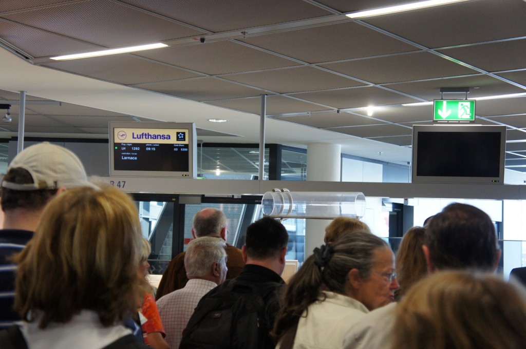 The image shows a group of people standing in line at an airport gate. Above the gate, there is a digital display board with the Lufthansa logo, indicating a flight to Larnaca with flight number LH 1292, scheduled to depart at 09:15. There is also an emergency exit sign with a green arrow pointing to the right. The ceiling has a modern design with recessed lighting. The people in the queue appear to be waiting to board the flight.
