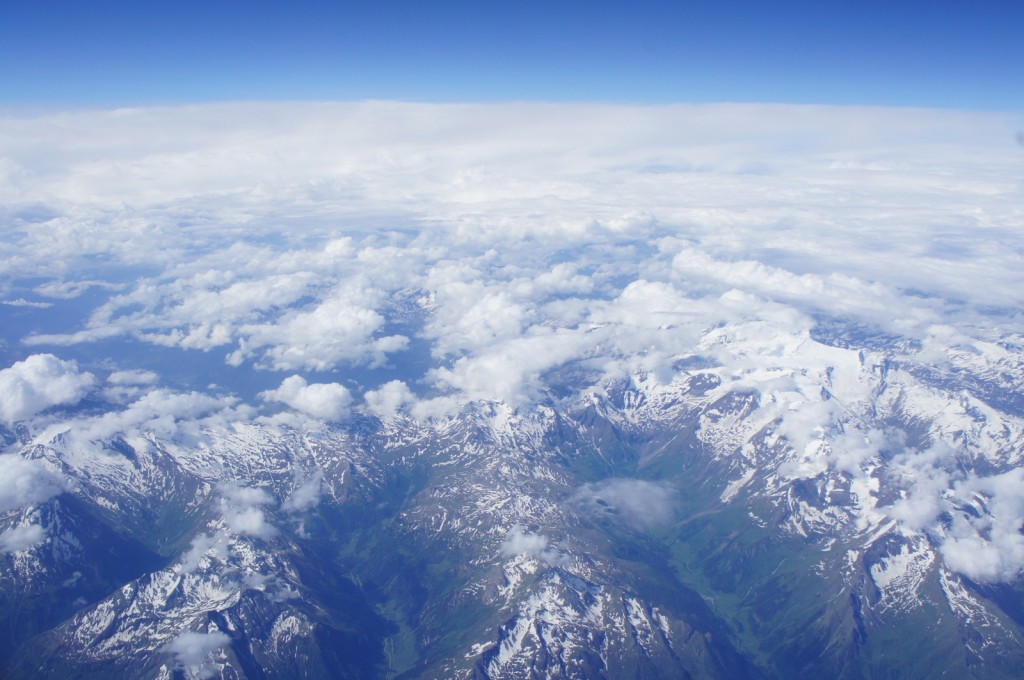 Aerial view of a mountain range with snow-capped peaks partially covered by clouds. The sky above is clear and blue, transitioning to a layer of white clouds in the distance. The landscape below shows a mix of rugged terrain and green valleys.