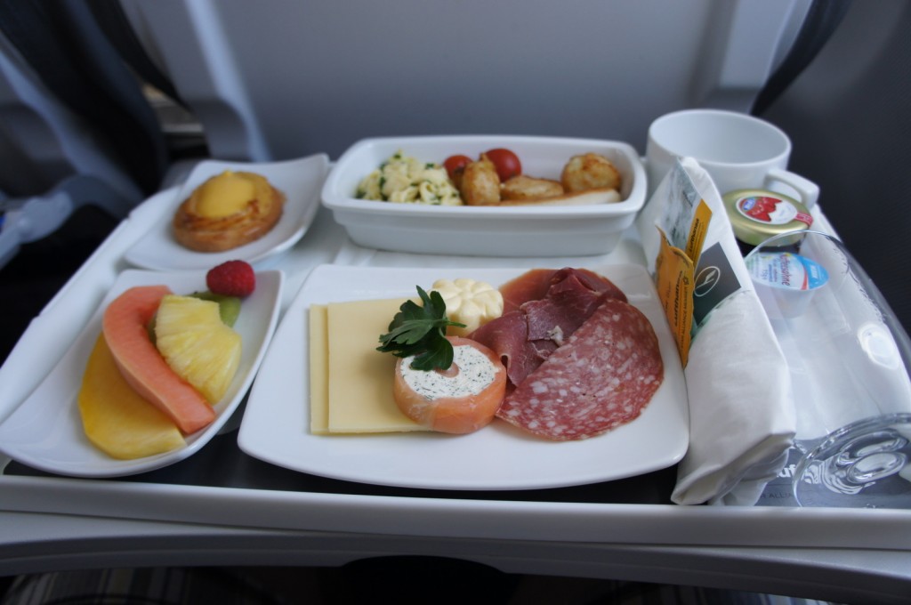 The image shows a meal served on an airplane tray. The tray includes a variety of food items: a plate with slices of cheese, salami, and other cold cuts, garnished with a sprig of parsley; a small dish with a pastry; a bowl with a salad containing pasta, cherry tomatoes, and roasted potatoes; a plate with slices of melon, pineapple, and a raspberry; a cup; a napkin with utensils; and small containers of butter and other condiments.