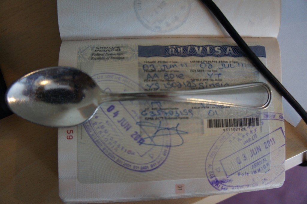 The image shows an open passport displaying a visa page with various stamps and handwritten details. A metal spoon is placed on top of the passport, partially covering some of the text and stamps. The visa is for the Federal Democratic Republic of Ethiopia, with visible dates and official stamps. The background includes a wooden surface and part of a black object, possibly a pen or cable, lying across the passport.