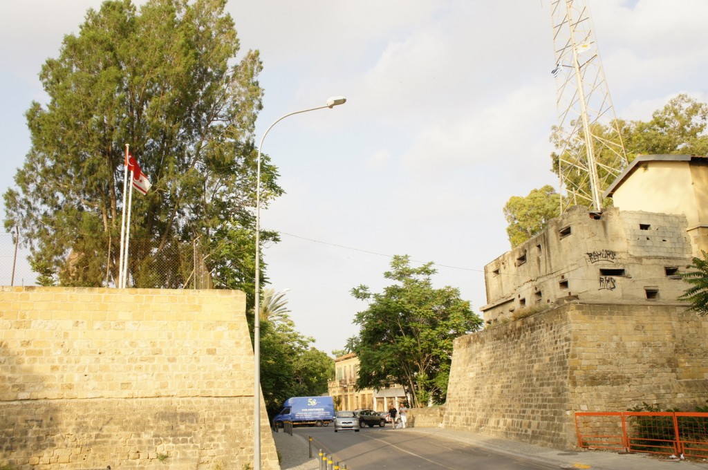 The image shows a street scene with a stone wall on the left side, topped with two flags. One flag is red with a white crescent and star, and the other is white with a red crescent and star. There is a tall tree behind the wall. On the right side, there is a large, old stone building with small windows and some graffiti on it. A tall metal structure, possibly a communication tower, is attached to the building. The street has a few cars and a blue van parked along it, and there are some people walking in the distance. The sky is partly cloudy.