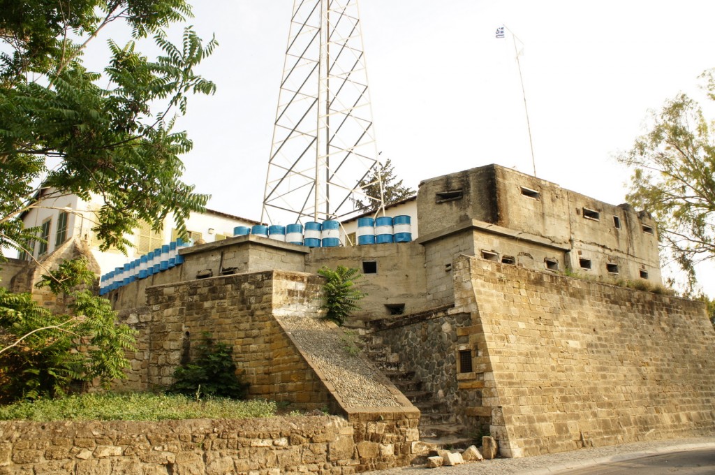 The image shows an old, fortified structure made of stone and concrete. The building has small, rectangular windows and appears to be a historical or military fortification. There are blue and white barrels lined up along the top of the structure. A tall metal tower and a flagpole with a flag are also visible on the roof. The surrounding area has some greenery, including trees and bushes. The overall scene suggests a mix of historical architecture and modern elements.