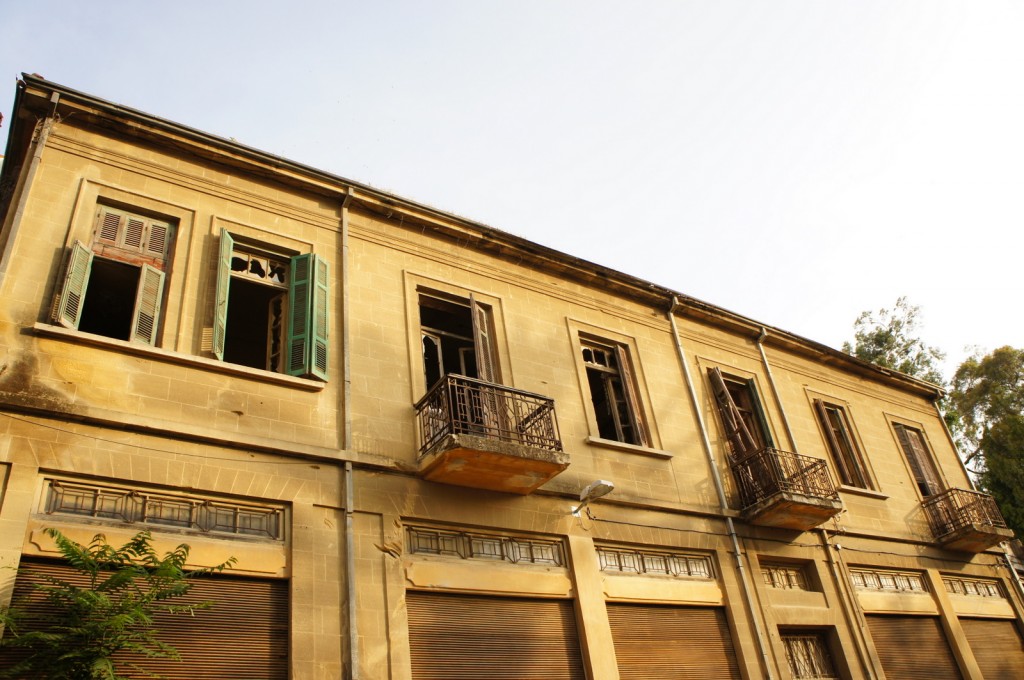 The image shows the facade of an old, abandoned two-story building. The building has a weathered appearance with peeling paint and broken windows. The upper floor features several windows, some with green shutters, and small balconies with metal railings. The lower floor has large, closed wooden shutters. The building is set against a clear sky, and there are some trees visible on the right side of the image.
