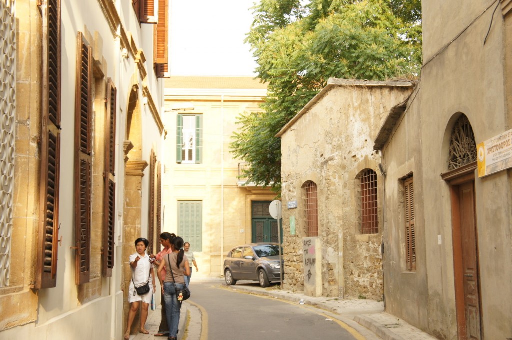 A narrow street in a Mediterranean town with old buildings on both sides. The buildings have wooden shutters and some have peeling paint. There are a few people standing on the left side of the street, engaged in conversation. A small car is parked further down the street. The scene is bathed in warm sunlight, and there are trees in the background.