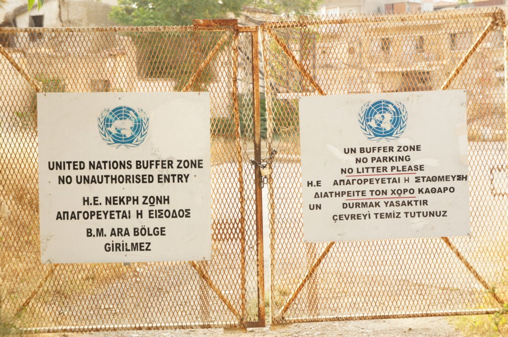 The image shows a metal gate with two signs attached to it. The signs display the United Nations emblem and contain text in multiple languages, including English, Greek, and Turkish. The left sign reads "UNITED NATIONS BUFFER ZONE NO UNAUTHORISED ENTRY" and the right sign reads "UN BUFFER ZONE NO PARKING NO LITTER PLEASE." The background appears to be an area with buildings and vegetation, indicating a restricted or controlled zone.