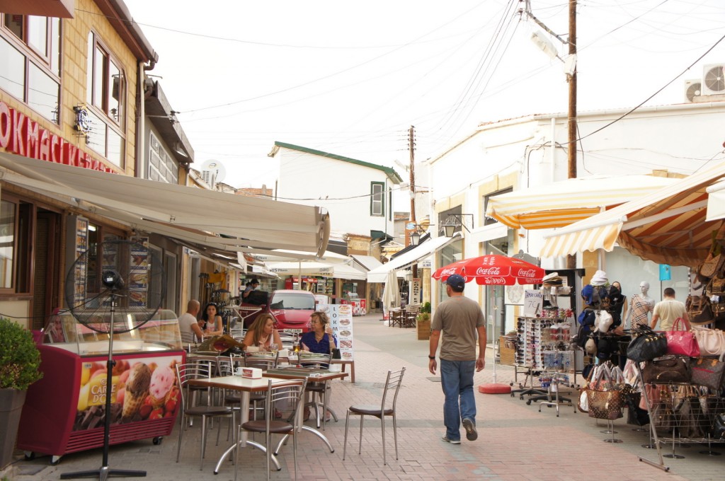 A narrow street lined with small shops and outdoor cafes. On the left side, there are tables and chairs set up outside a cafe, with a few people sitting and eating. An ice cream freezer with a colorful advertisement is also visible. On the right side, there are various stalls selling items like bags, hats, and sunglasses. A man is walking down the middle of the street, and there are several awnings and umbrellas providing shade. The street has a relaxed, casual atmosphere.