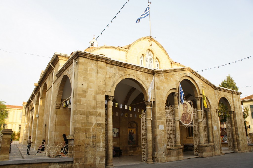 The image shows a historic stone church with arched entrances and a Greek flag flying on top. The building has a traditional architectural style with multiple arches and columns. There are also other flags and religious icons displayed near the entrance. Some people are seen sitting and standing around the church. The sky is clear, indicating a sunny day.