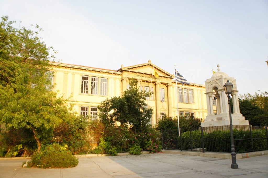 The image shows a neoclassical building with a symmetrical facade, featuring tall windows and a central pediment supported by columns. In front of the building, there is a well-maintained garden with various trees and shrubs. To the right of the building, there is a white stone monument with an arched structure and a flagpole displaying a flag. The scene is well-lit, suggesting it is daytime.