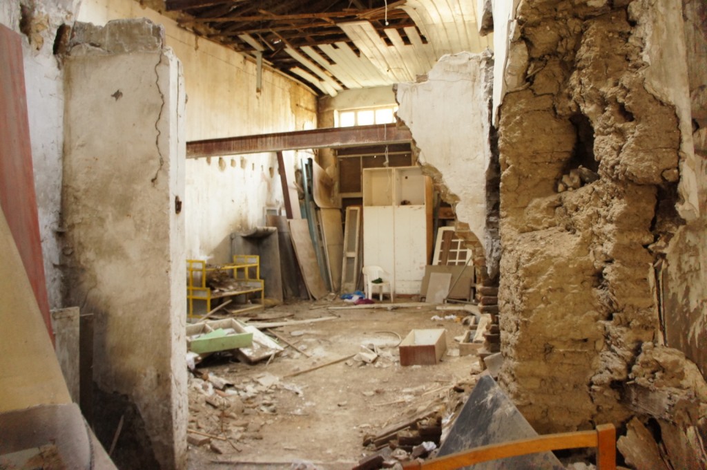 The image shows the interior of a dilapidated and abandoned building. The room is in a state of disrepair, with crumbling walls, exposed wooden beams, and debris scattered across the floor. There are various construction materials and old furniture pieces, such as shelves and a chair, strewn around the space. The ceiling has large gaps, and the overall appearance suggests that the building has been neglected for a long time.