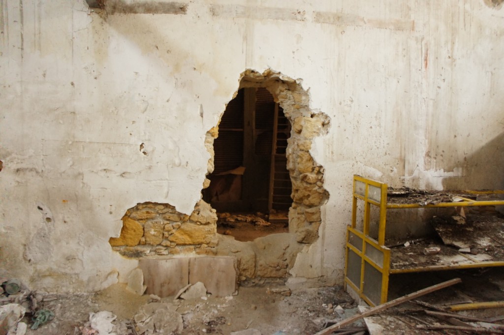 The image shows an interior scene of a dilapidated building. There is a large hole in a white, worn-out wall, revealing a dark space behind it. The wall has visible cracks and peeling paint. To the right of the hole, there is a yellow metal shelving unit with debris and dirt on it. The floor is covered with rubble and broken pieces of the wall. The overall atmosphere is one of decay and abandonment.